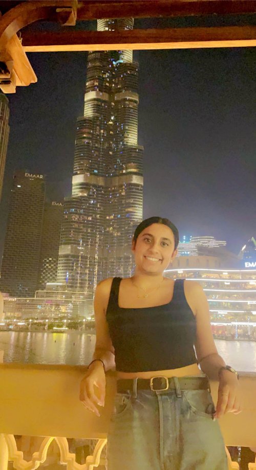 A young woman in a black top with dark features leans on a railing around a body of water and smiles at the camera. In the background is the Burj Khalifa and other skyscrapers. It is night.
