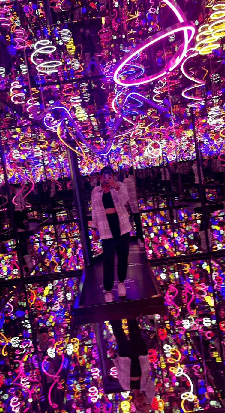 A young woman stands in a dark room lined with mirrors reflecting colorful neon curls and squiggles. She holds a phone in front of her face to take the photo.